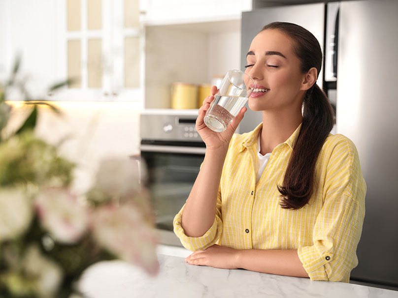 A woman with brown hair and a yellow blouse drinking a glass of water in her kitchen A woman with brown hair and a yellow blouse drinking a glass of water in her kitchen